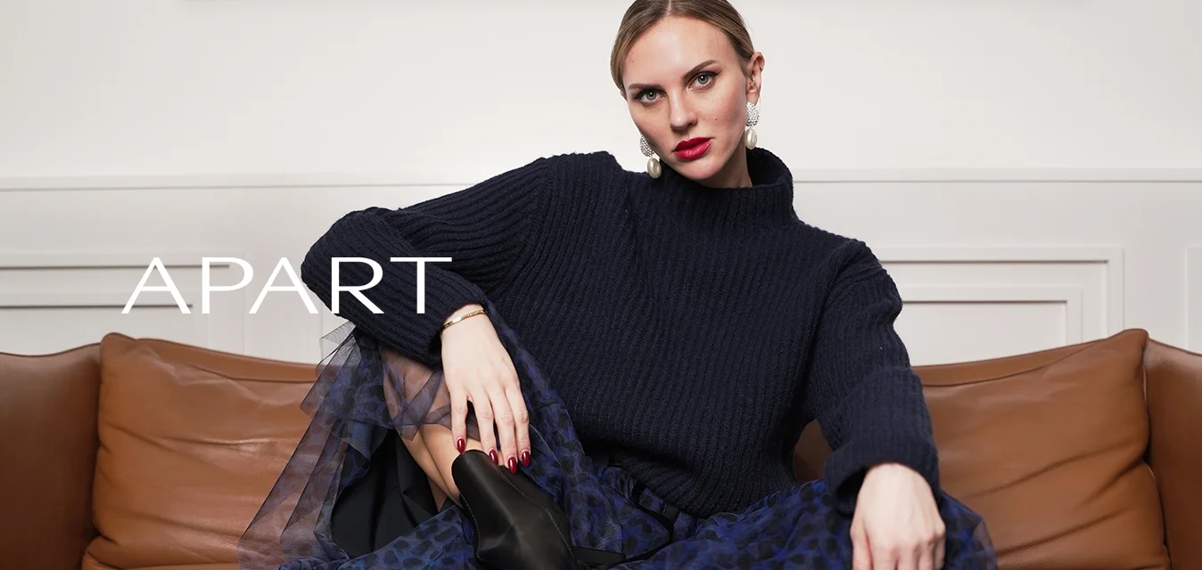 Maglione a coste navy, indossato sopra una gonna in tulle blu trasparente con un rivestimento interno nero. Stivaletti neri alla caviglia. Sfondo con divano in pelle marrone.