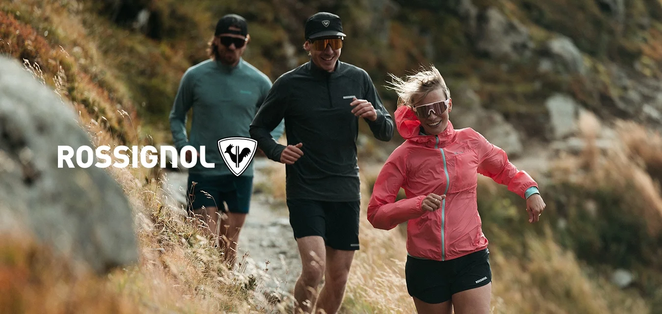 Tre corridori in tenuta atletica scendono lungo un sentiero roccioso circondato da erba e terreno ripido.