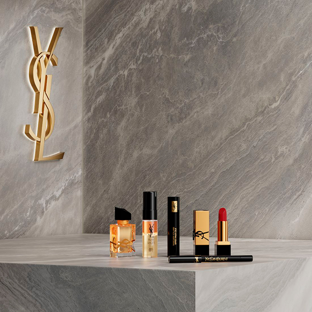 Una collezione di prodotti di bellezza YSL, inclusi profumo, mascara, rossetto e eyeliner, &egrave; elegantemente disposta su una superficie di marmo grigio chiaro con un grande logo YSL illuminato sulla parete di marmo abbinata sullo sfondo.