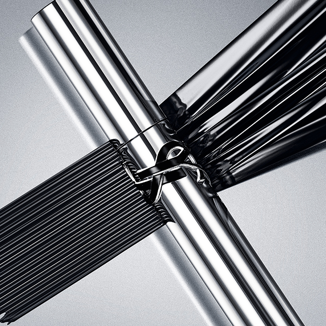 Due tubetti di mascara YSL argentati metallici, adornati con il logo YSL, sono incrociati su una superficie grigio chiaro lucida, circondati da ricchi tratti di mascara nero che creano un motivo dinamico.