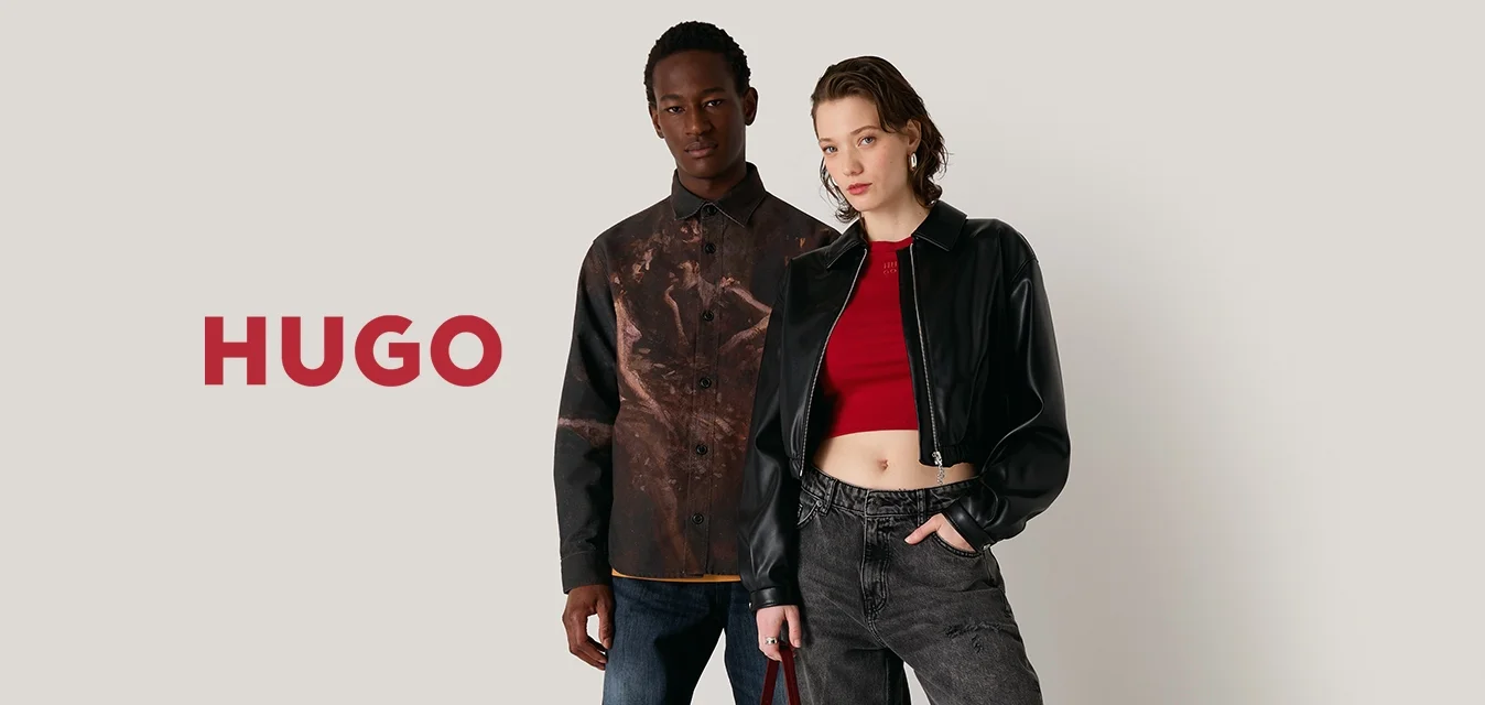 Due modelli si trovano di fronte a uno sfondo neutro. Uno indossa una camicia a fantasia marrone e jeans blu, l'altro una giacca cropped nera e pantaloni grigi.