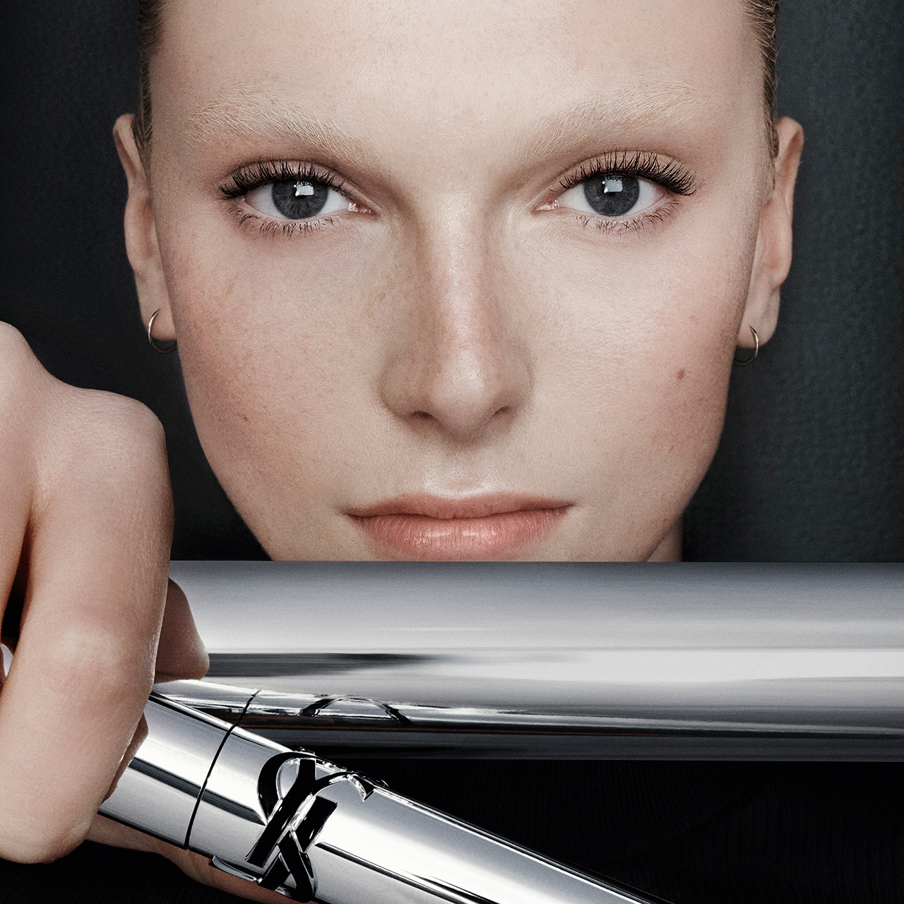 Un primo piano del viso di una modella con capelli chiari e occhi azzurri, che guarda direttamente in avanti, mentre la sua mano tiene un tubetto di mascara YSL argentato, nascondendo parzialmente la parte inferiore del suo viso.