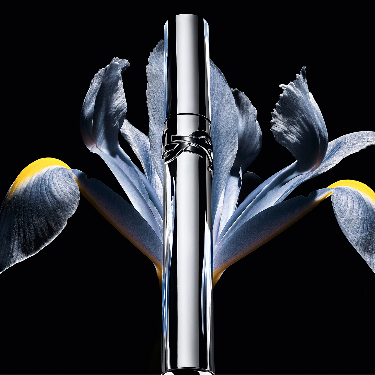 Un tubetto di mascara YSL argentato, con il logo del marchio, si erge verticalmente tra i delicati petali azzurri di un fiore di iris con accenti gialli, il tutto su uno sfondo scuro.