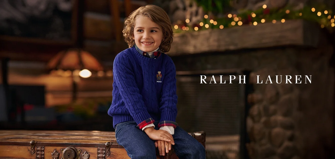 Bambino sorridente con capelli ricci, vestito con un maglione azzurro a trecce e jeans, seduto su un antico baule di legno in una stanza accogliente con camino in pietra.