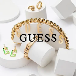 Gioielli Guess in oro, tra cui un bracciale a catena, due anelli e orecchini verdi, esposti su forme geometriche bianche su sfondo bianco.