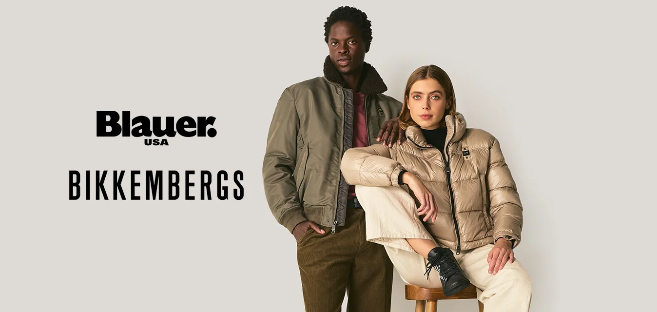 Due adulti indossano giacche calde: uno in piedi con un bomber oliva e pantaloni marroni, l'altro seduto su uno sgabello con un piumino beige e pantaloni crema.