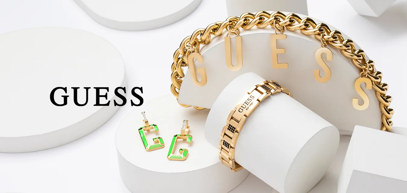 Gioielli Guess in oro, tra cui un bracciale a catena, due anelli e orecchini verdi, esposti su forme geometriche bianche su sfondo bianco.