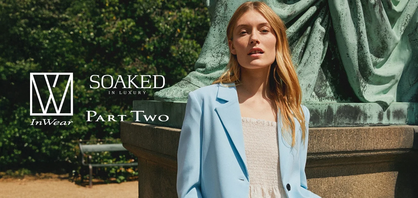 Donna con i capelli lunghi biondi che indossa un blazer blu chiaro e un vestito bianco, si trova vicino a un grande piedistallo verde all'aperto.