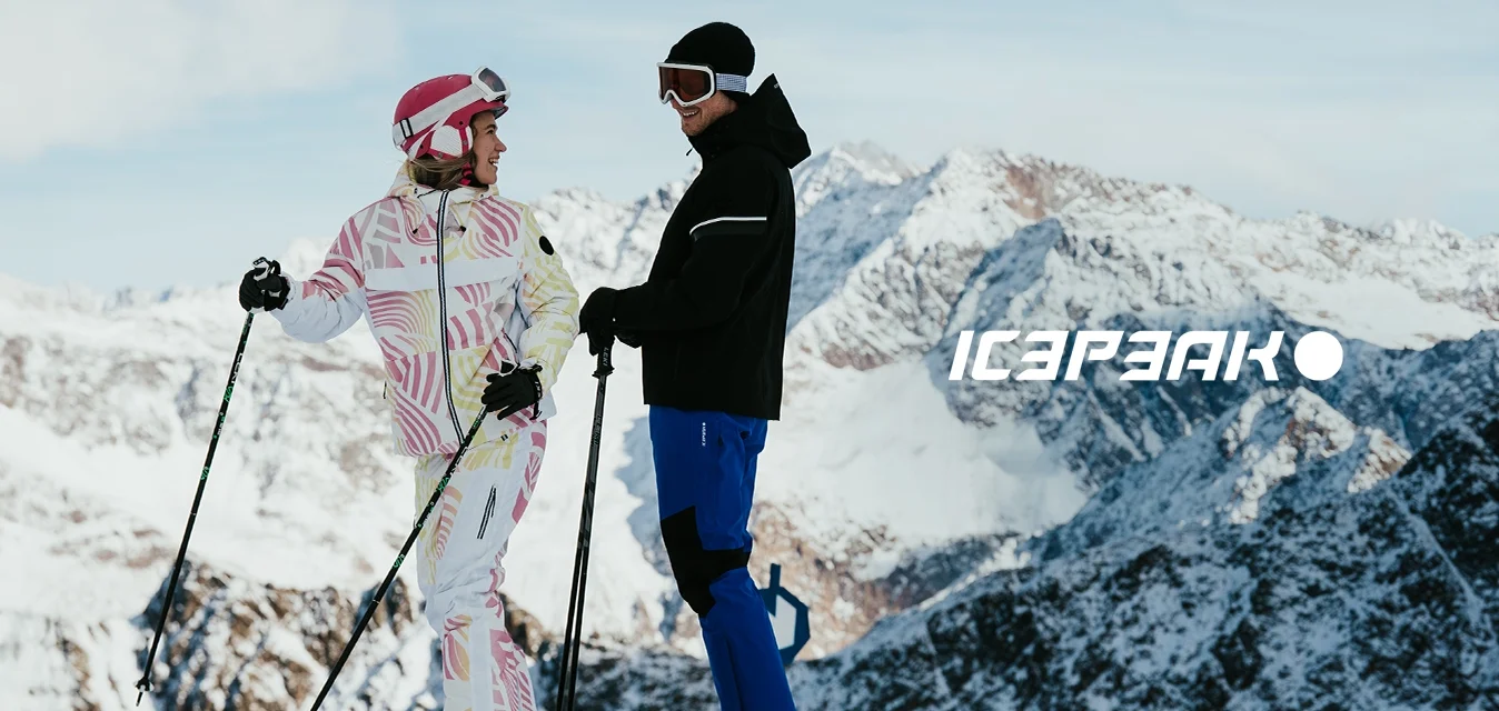 Due snowboarder in piedi sulla neve, uno con una tuta rosa e bianca a motivi, l'altro con una giacca nera e pantaloni blu. Sullo sfondo, montagne innevate.