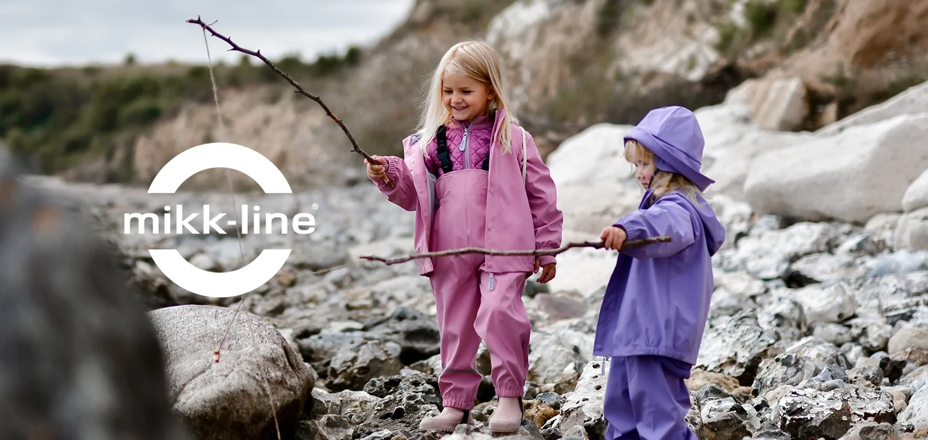 Due bambini su una costa rocciosa, vestiti con tute impermeabili rosa e viola, ciascuno con in mano un bastone. Rocce texturizzate e una collina erbosa sullo sfondo.