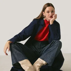 Donna seduta per terra con le gambe divaricate, indossa un maglione rosso, un cappotto blu navy, pantaloni scuri e stivali beige su sfondo chiaro.