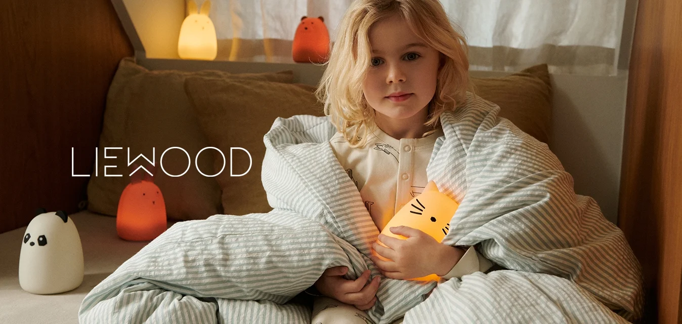 Un bambino avvolto in una coperta a righe blu chiaro tiene in mano una lampada a forma di gatto che emette una luce soffusa. Si possono vedere morbidi cuscini e lampade a forma di animali in colori caldi.