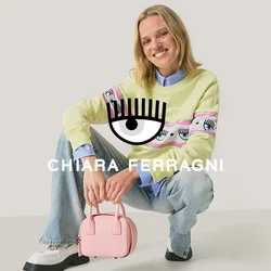 Felpa verde chiaro con grafica a occhio, indossata sopra una camicia blu; abbinata a jeans di denim chiaro e una piccola borsa rosa. Scarpe nere.