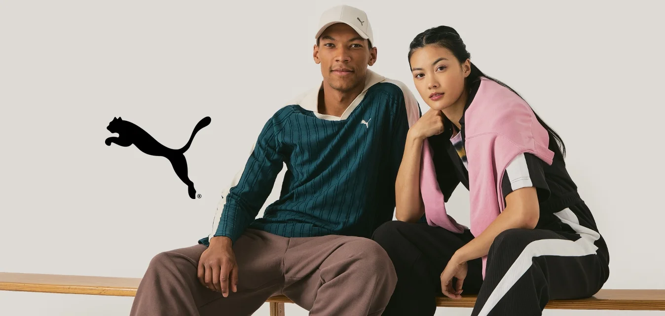 Due modelli siedono su una panchina di legno. L'uomo indossa una maglietta a righe verdi e pantaloni marroni, mentre la donna ha un tuta nera con dettagli rosa.