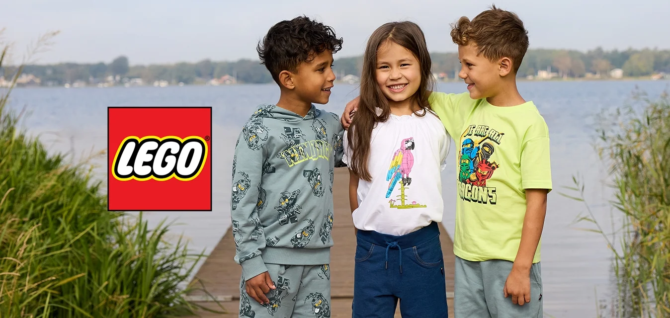 Tre bambini sono in piedi su un sentiero di legno vicino all'acqua. Indossano outfit casual: una felpa verde con stampe, una maglietta bianca con un design di pappagallo e pantaloni corti vivaci.