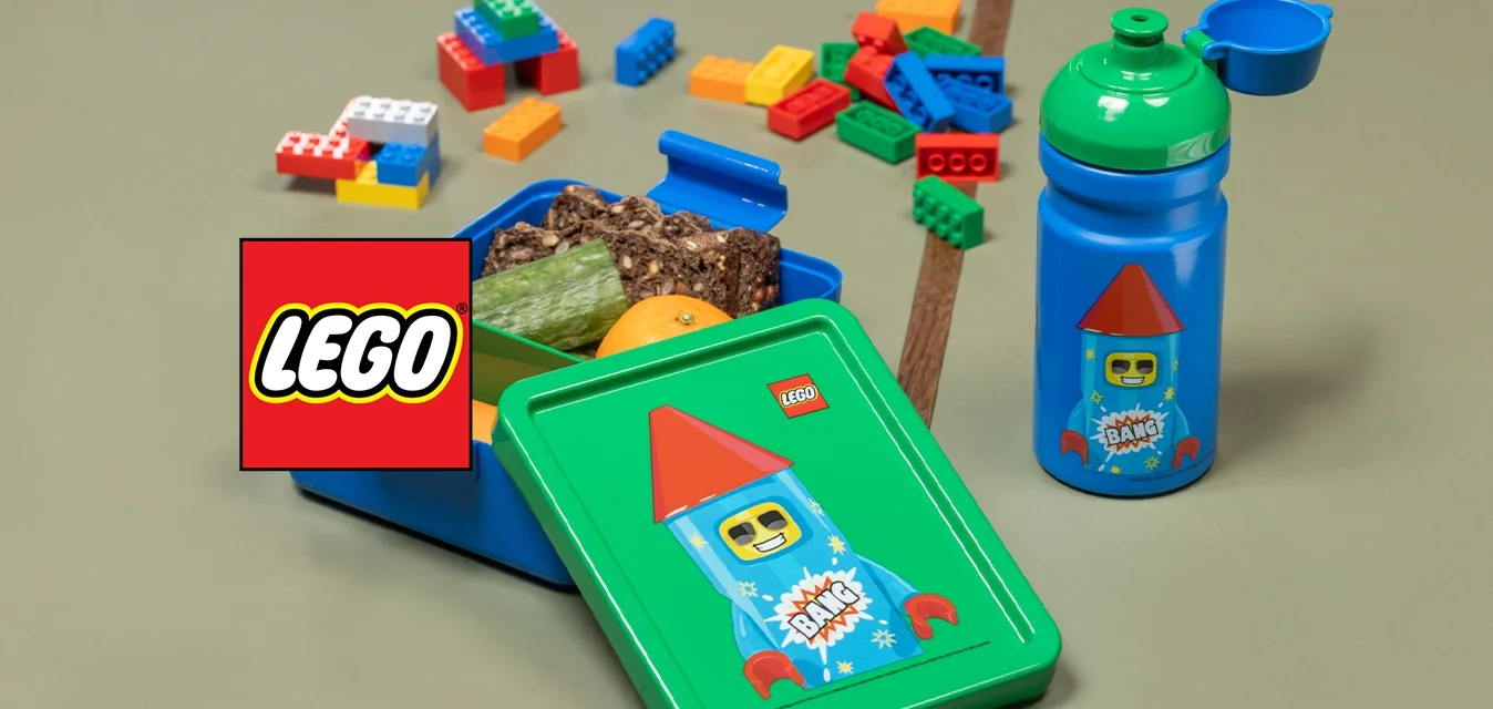 Lunchbox blu a tema LEGO con coperchio verde, riempita di snack e un'arancia, accanto a una bottiglia blu abbinata con tappo verde, su un tavolo.