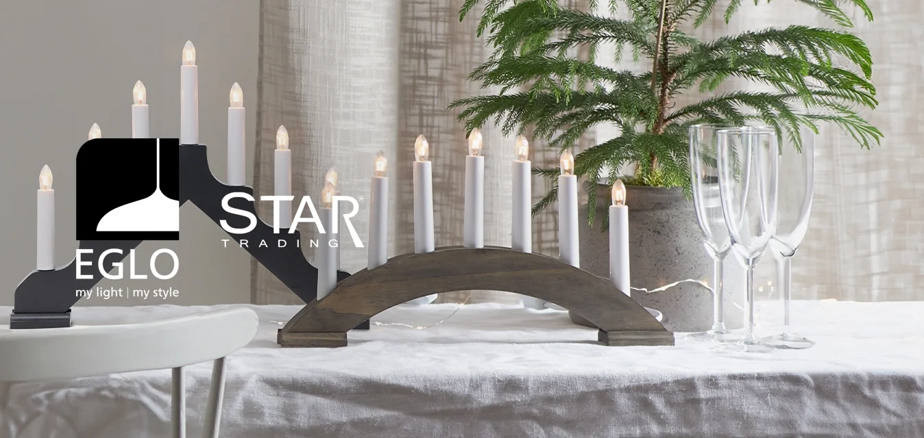 Due portacandele, uno nero con una forma ondulata e uno ad arco in legno, con candele bianche. Sullo sfondo, una pianta in vaso e bicchieri su una tovaglia bianca.