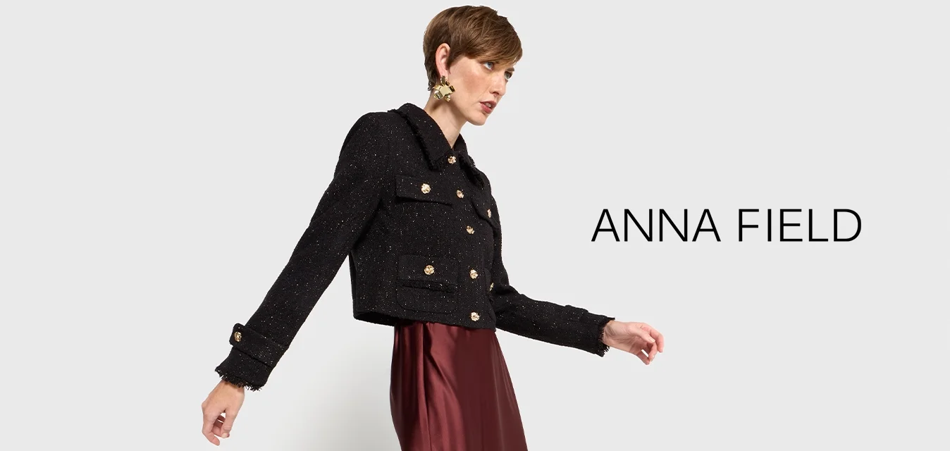 Giacca nera corta con bottoni dorati, abbinata a una gonna maxi in satin borgogna, completata da stivali neri al ginocchio. Texture liscia.