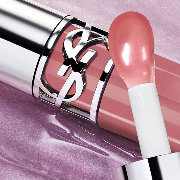 Un primo piano di un lucidalabbra YSL aperto, con l'applicatore rivestito di un gloss rosa scintillante, che emerge dal tubo trasparente su uno sfondo rosa sfocato e riflettente.