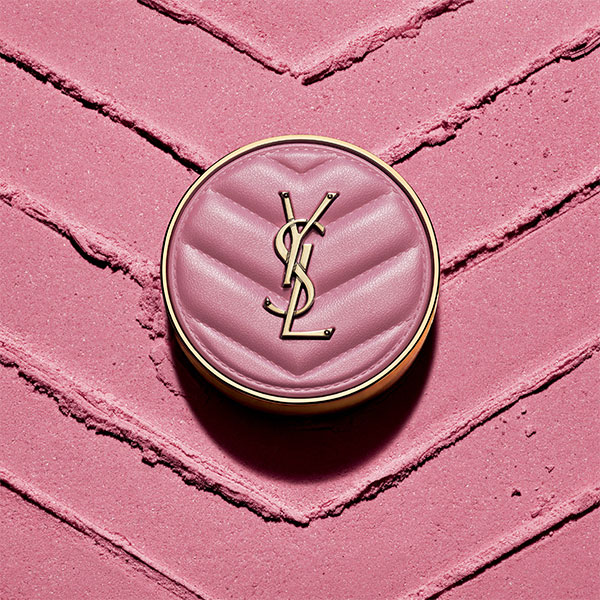 Un fondotinta cushion YSL circolare con un coperchio rosa trapuntato e un logo YSL dorato &egrave; centrato su uno sfondo di polvere rosa testurizzata con motivi a spina di pesce.