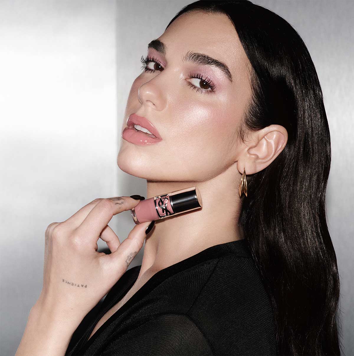 La cantante Dua Lipa, con lunghi capelli scuri, ombretto rosa delicato e labbra rosa chiaro, tiene elegantemente un prodotto labbra YSL vicino al collo, guardando direttamente lo spettatore.