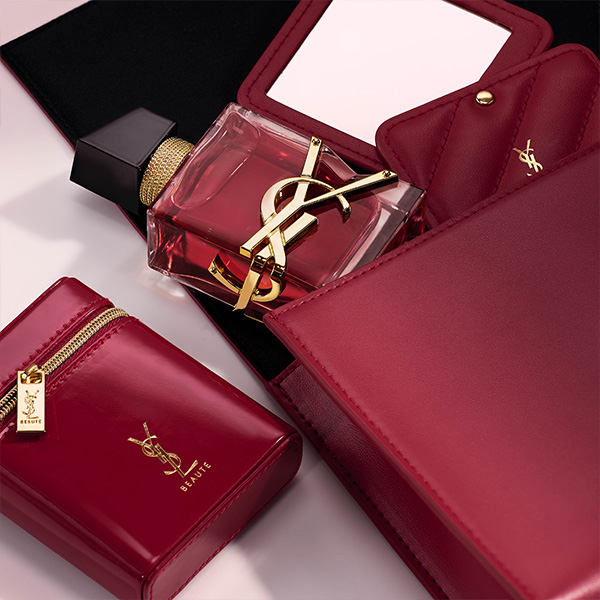 Un flacone di profumo YSL Libre con liquido rosso &egrave; esposto tra lussuosi accessori in pelle rossa, inclusi un compatto con specchio e una pochette con cerniera.