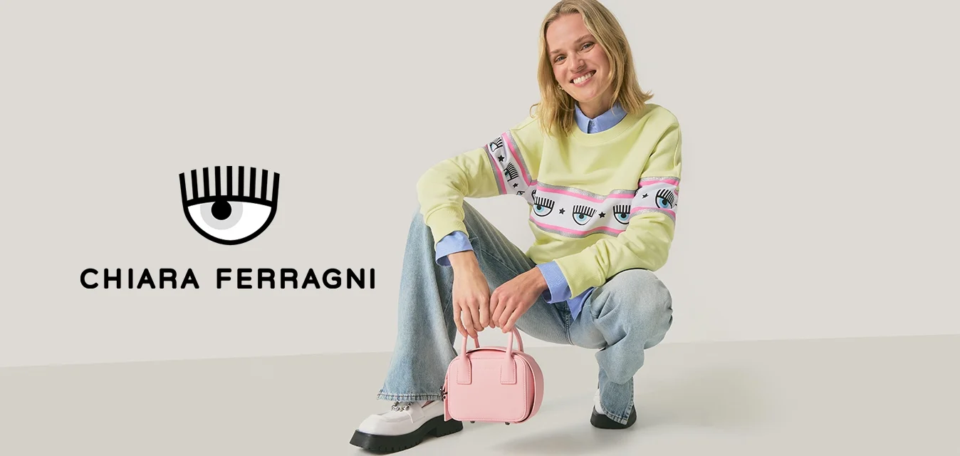 Felpa verde chiaro con grafica a occhio, indossata sopra una camicia blu; abbinata a jeans di denim chiaro e una piccola borsa rosa. Scarpe nere.