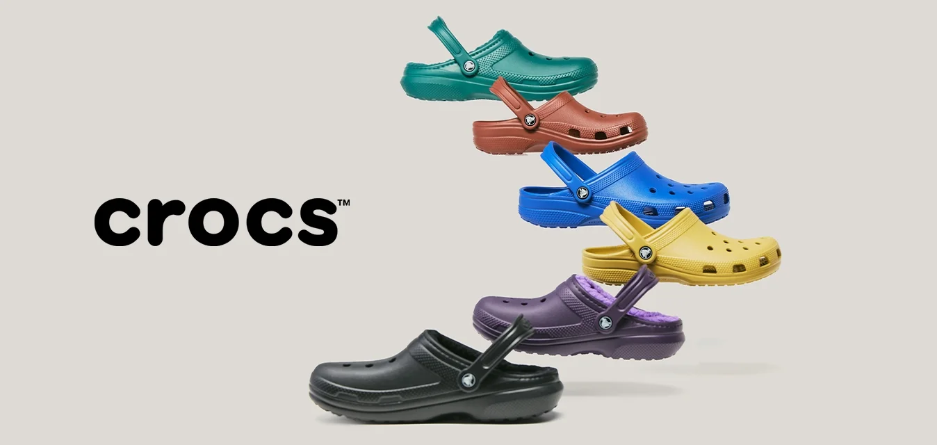 Sei colori vivaci di clogs Crocs in nero, viola, giallo, blu, rosso e verde, disposti in una formazione verticale sfalsata su uno sfondo chiaro.