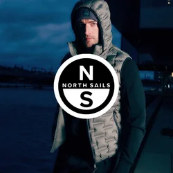 Gilet imbottito grigio con cappuccio, caratterizzato da un design trapuntato e una toppa con logo, indossato sopra una maglia nera a maniche lunghe, sullo sfondo di un lungomare.