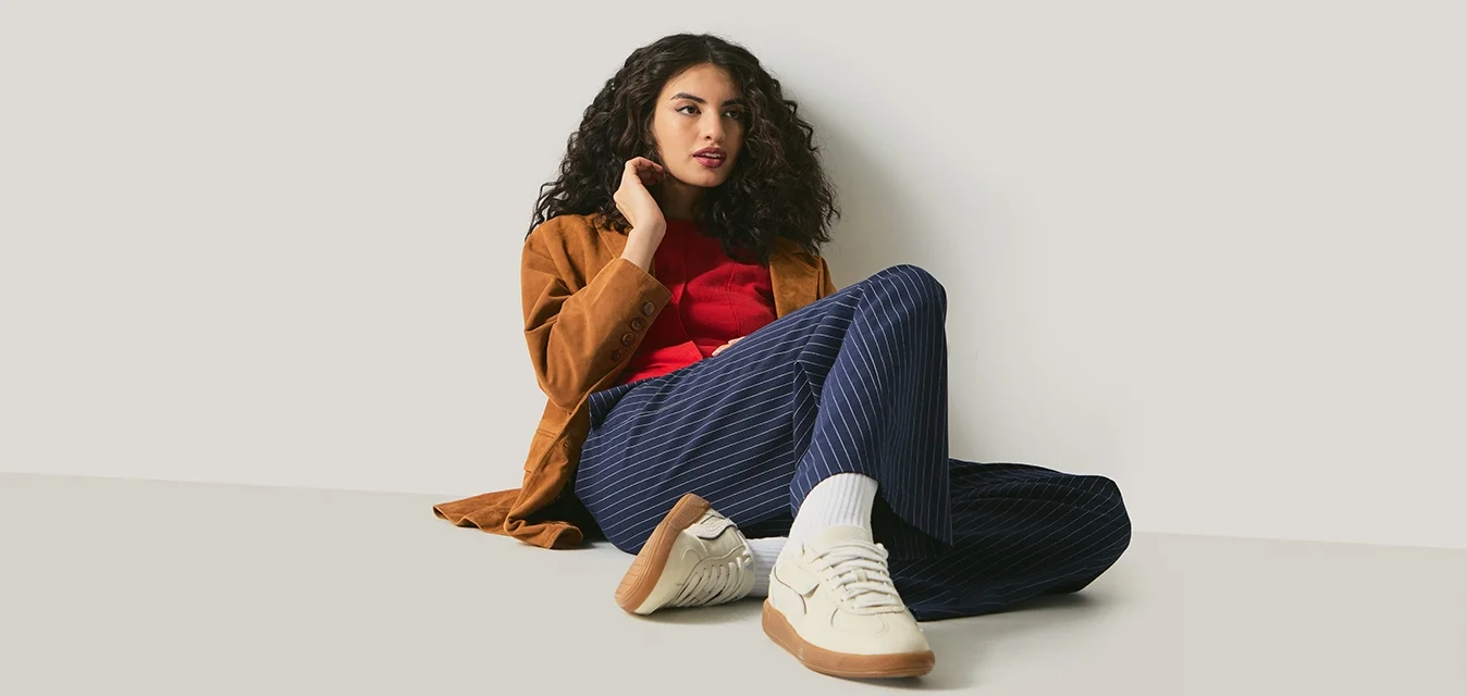 Giovane donna con capelli ricci, che indossa una giacca marrone, una camicia rossa, pantaloni blu navy a righe sottili e scarpe da ginnastica bianche, seduta sul pavimento contro una parete bianca.