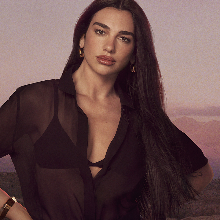 La cantante Dua Lipa, con lunghi capelli scuri, indossa una camicia nera trasparente sopra un reggiseno e posa con sicurezza contro uno sfondo montuoso morbido nei toni del viola e del rosa.
