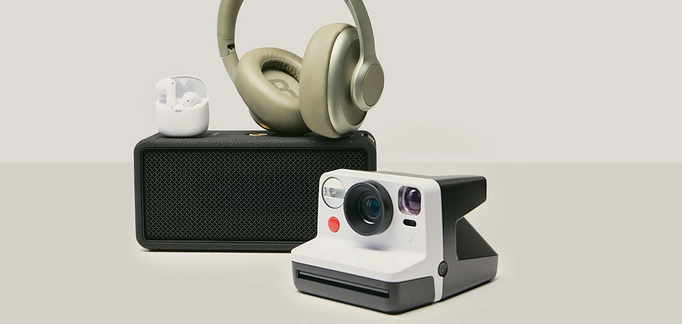 Cuffie wireless appoggiate su un altoparlante nero, auricolari wireless bianchi in una custodia e una fotocamera istantanea in stile vintage su una superficie neutra.