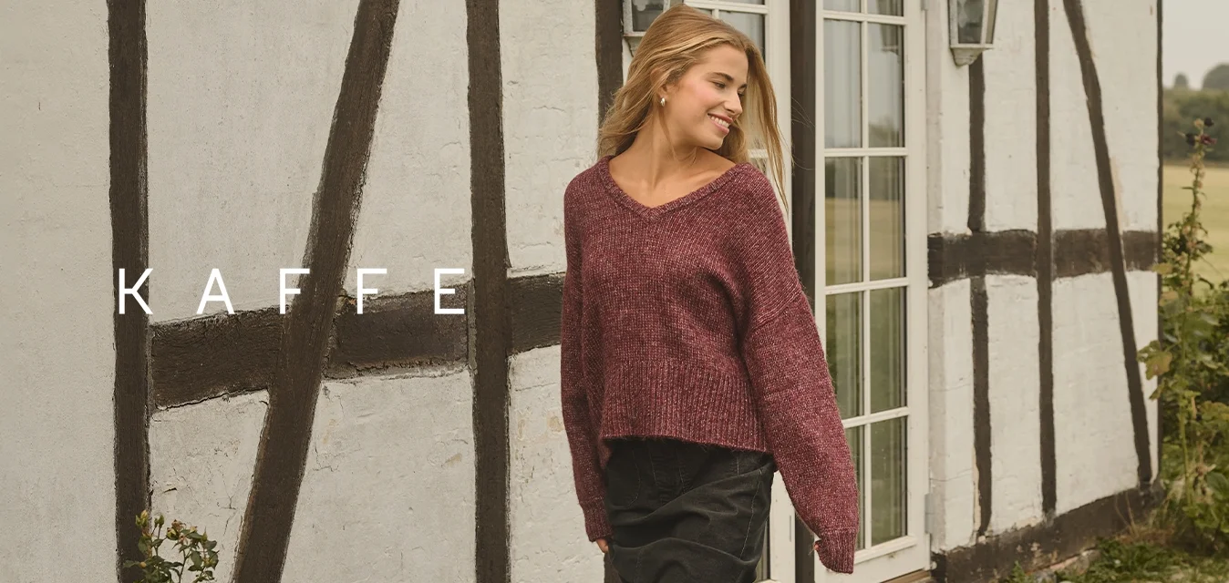 Giovane donna in un maglione bordeaux e stivali neri che cammina su un sentiero di ciottoli accanto a un cottage con tetto di paglia e fiori bianchi nei dintorni.