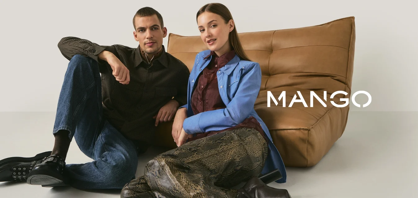 Giovane uomo e donna seduti sul pavimento appoggiati a un divano in pelle color cuoio, vestiti con abiti casual e alla moda contro uno sfondo semplice.