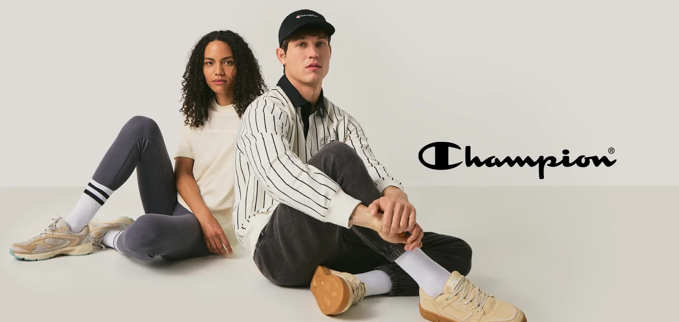 Un uomo e una donna sono seduti sul pavimento schiena contro schiena, entrambi indossano abbigliamento casual e sneaker beige, su uno sfondo chiaro e neutro.