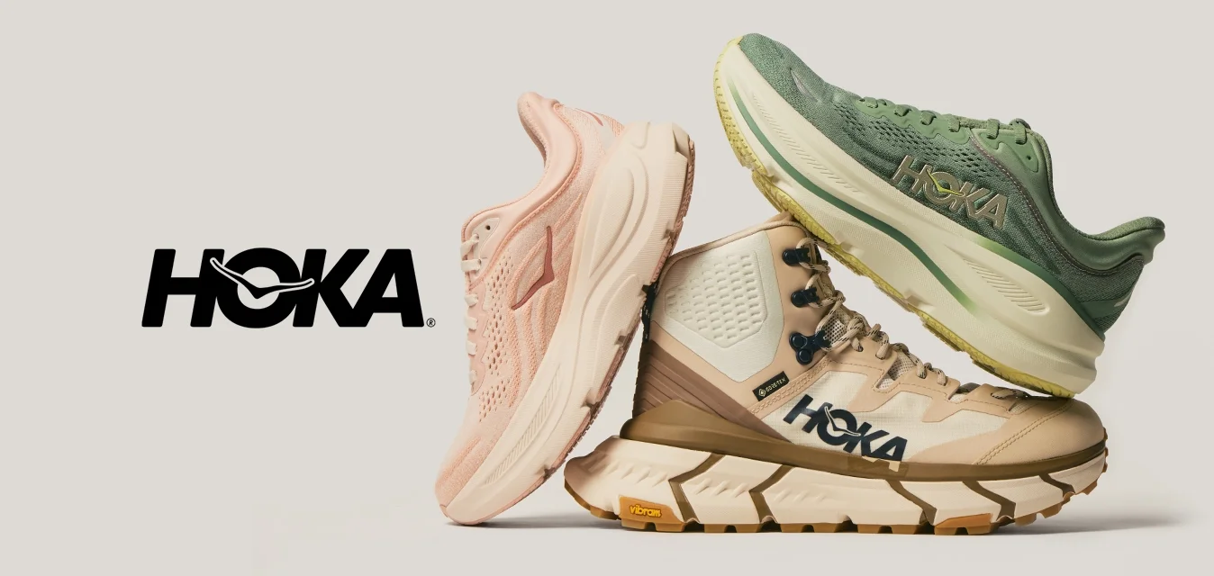 Tre paia di scarpe da corsa HOKA impilate, in rosa, un boot da escursionismo beige e verde, viste di lato su uno sfondo neutro chiaro.