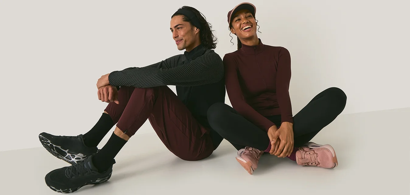 Due persone siedono su uno sfondo chiaro. L'uomo indossa una maglietta nera a trama, pantaloni bordeaux e scarpe nere. La donna indossa una giacca bordeaux con zip, leggings neri e scarpe rosa.