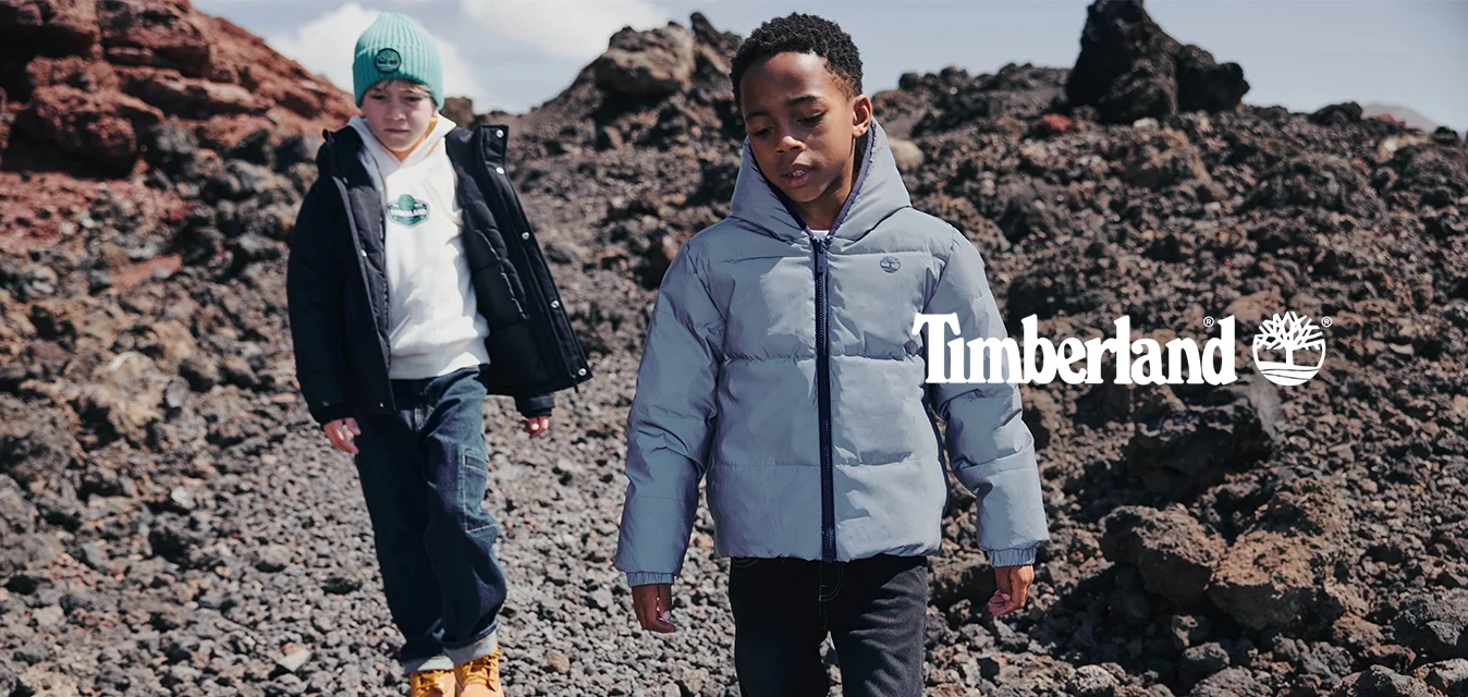 Due ragazzi con giacche invernali che camminano su un terreno roccioso con rocce vulcaniche sotto un cielo parzialmente nuvoloso.