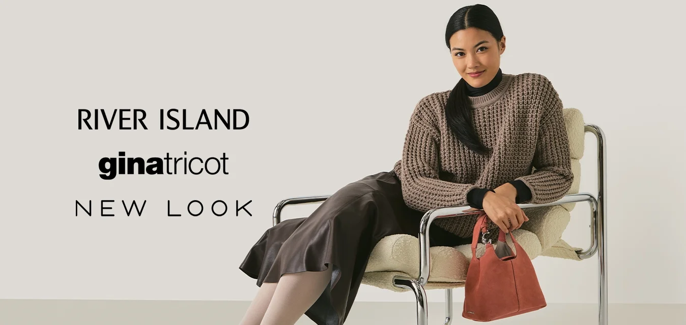 Donna con un maglione di lana marrone e una gonna di pelle, seduta su una moderna sedia in cromo e tessuto, che tiene una piccola borsa rossa, sorridendo verso la fotocamera.