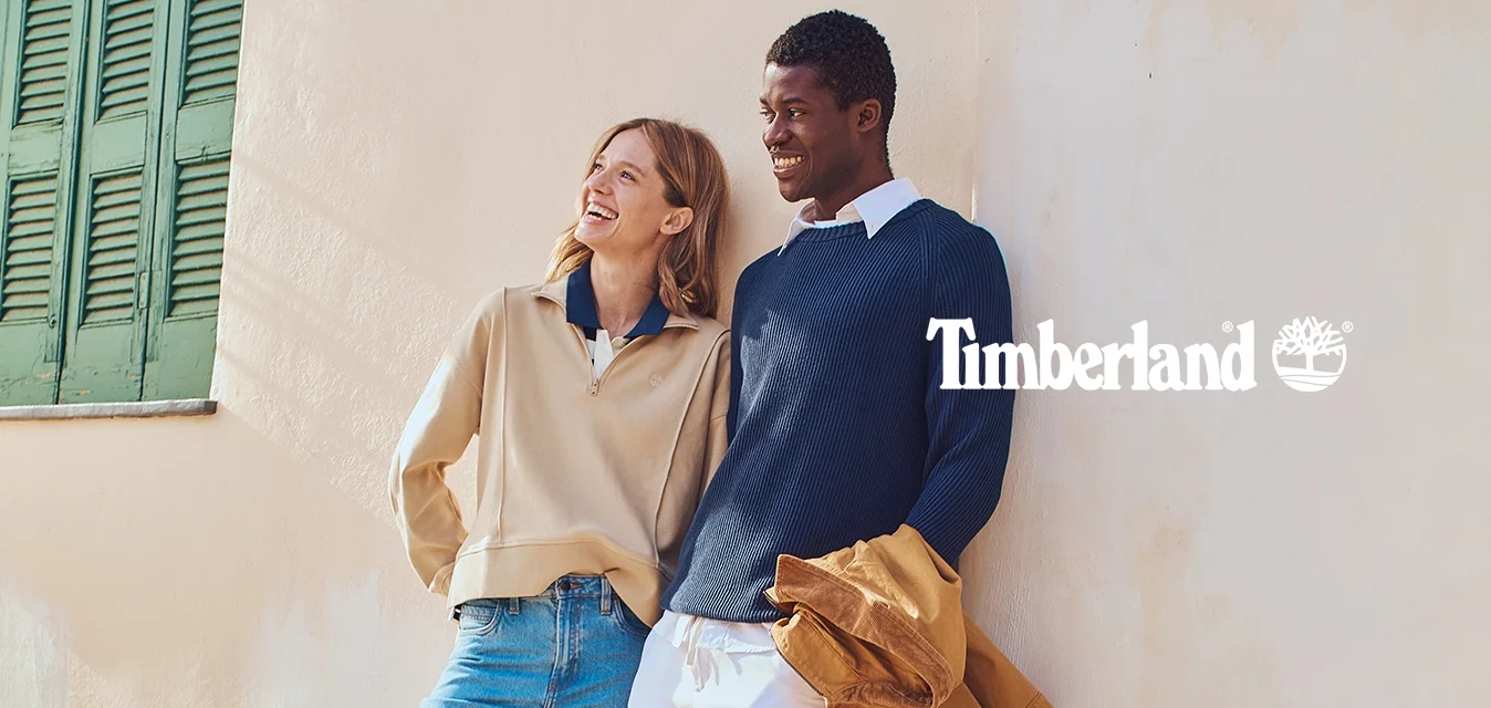 Maglione beige con zip, colletto blu; maglione blu navy a coste; jeans azzurri; scarpe marroni; pantaloni bianchi; giacca marrone; parete testurizzata; persiane verdi.