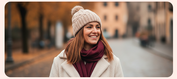 Donna sorridente con cappotto chiaro, sciarpa bordeaux e berretto di lana beige passeggia in una strada autunnale o invernale, con sfondo sfocato e alberi dalle foglie arancioni.