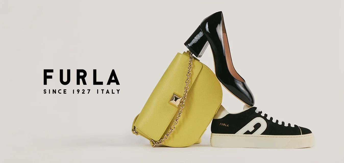 Scarpa con tacco alto in pelle verniciata nera in equilibrio su una borsa gialla, accanto a una sneaker casual bianca e nera su sfondo bianco semplice.