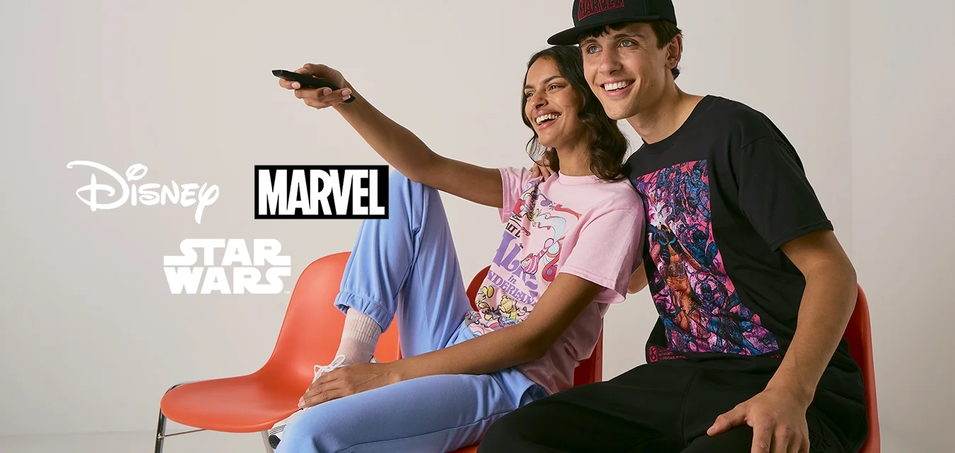 Due persone sedute su sedie di plastica arancioni, che indossano magliette grafiche; una rosa con un cartone animato e l'altra nera con arte colorata.