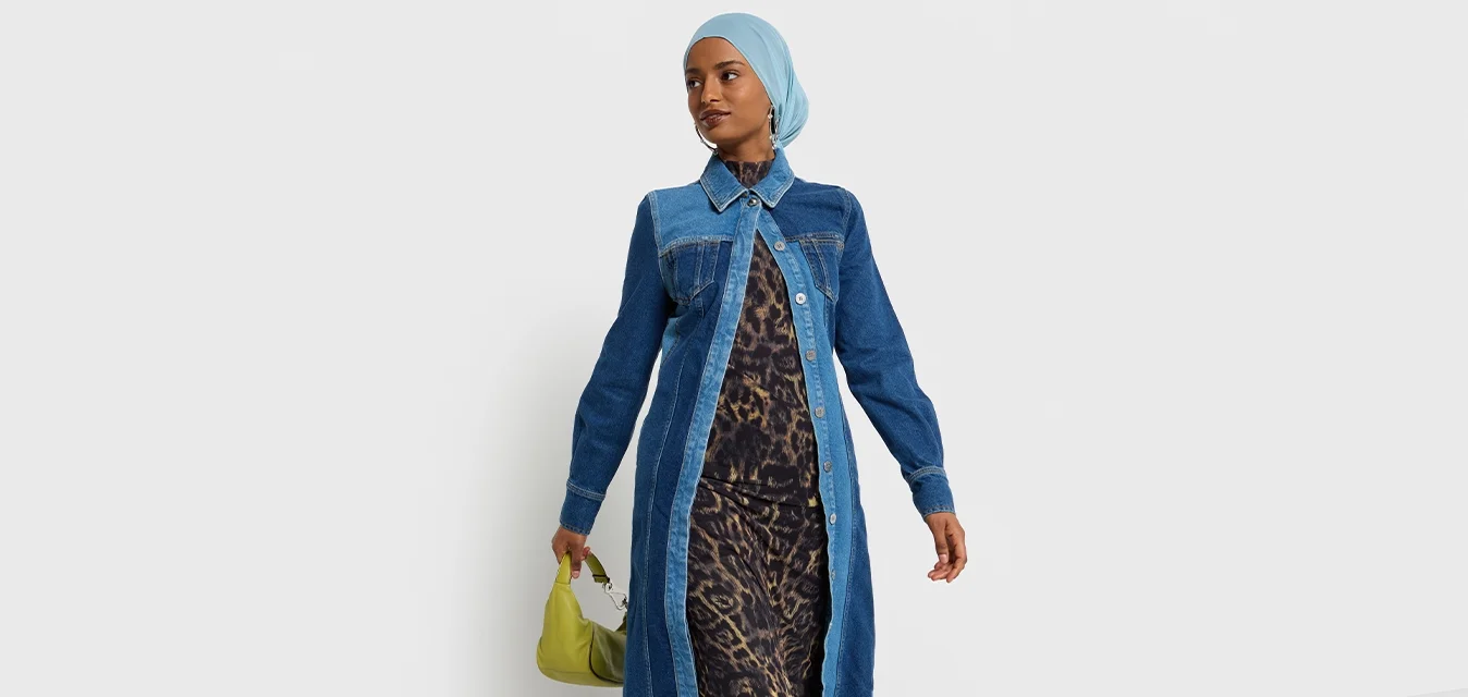 Cappotto lungo in denim con colletto, pannelli blu a contrasto e chiusura con bottoni. Sotto, un vestito con stampa leopardata e borsa verde.