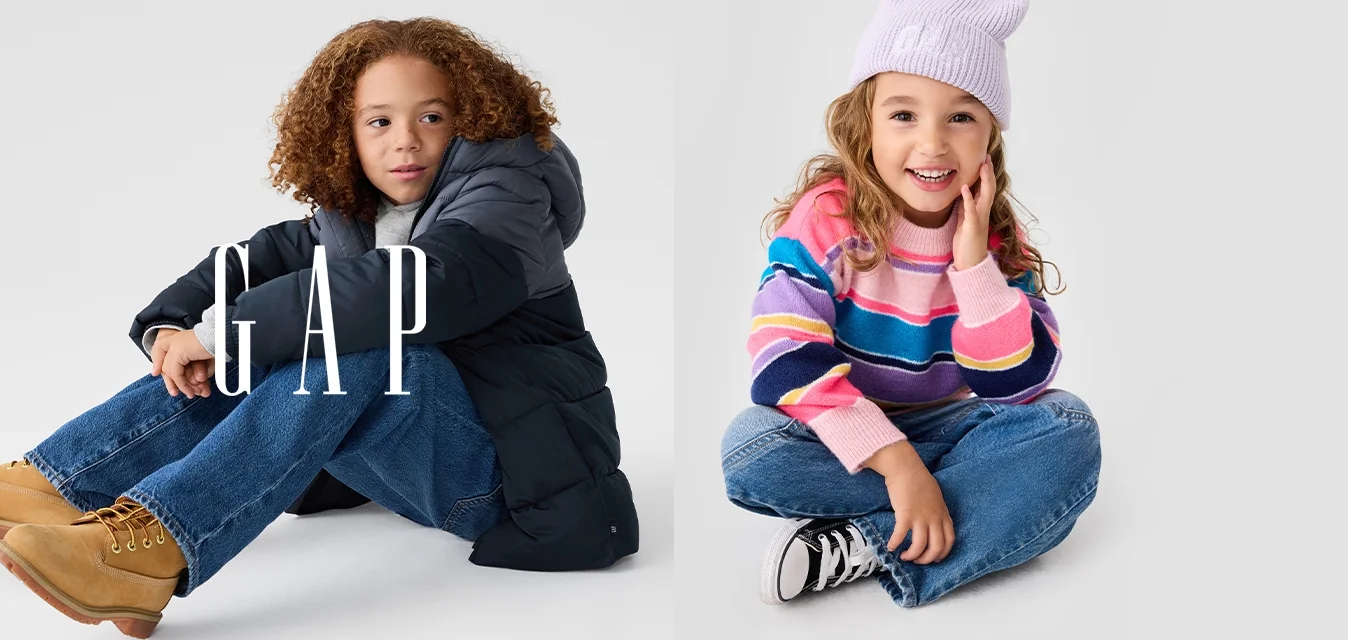 Due bambini seduti su uno sfondo neutro. Uno indossa una giacca imbottita scura e jeans azzurri; l'altro porta un maglione a righe e un berretto lavanda.