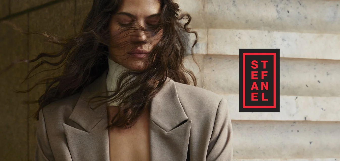 Blazer beige con vestibilità rilassata, morbido al tatto e con rever affilati, abbinato a un top bianco a coste. I capelli mossi e scompigliati dal vento coprono parzialmente il viso.