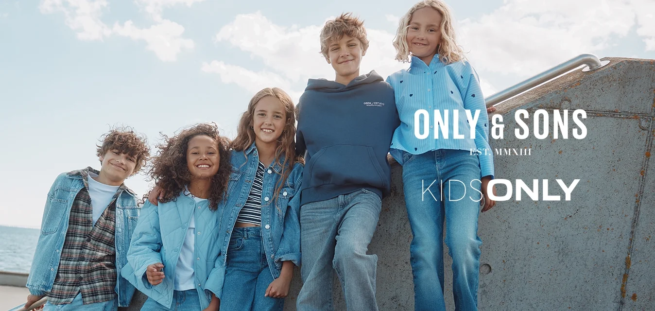 Cinque bambini vestiti con abiti casual in denim e giacche sorridono insieme su gradini di cemento all'aperto vicino al mare sotto un cielo parzialmente nuvoloso.