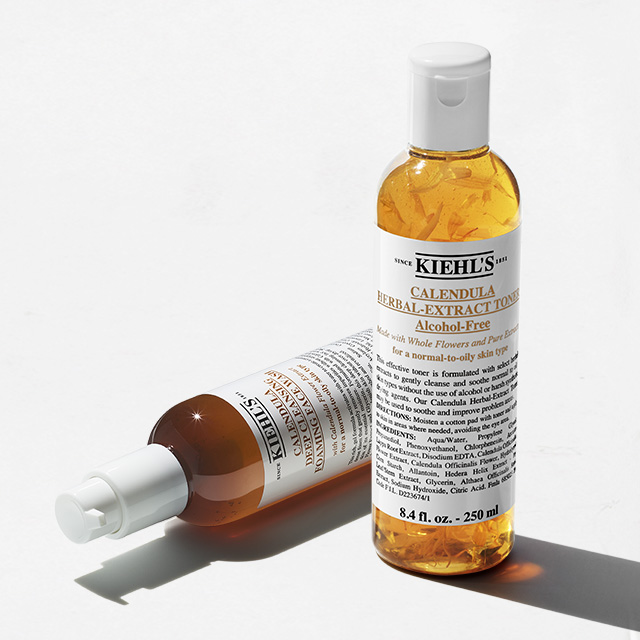 Flaconi di Calendula Herbal-Extract Toner e Calendula Foaming Wash  di Kiehl&rsquo;s, uno in piedi e uno sdraiato, su fondo bianco, con petali di calendula visibili nel liquido.