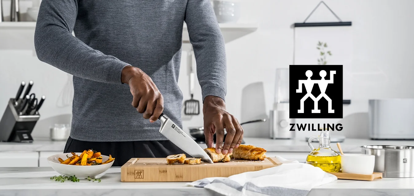 Coltello da chef con lama in acciaio inossidabile, tagliere in legno, pollo affettato, maglione grigio, asciugamano bianco con striscia grigia, allestimento da cucina.