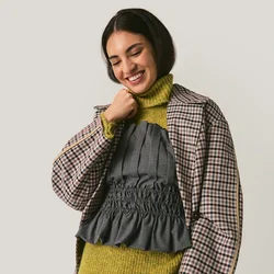 Giovane donna sorridente, indossa un blazer a quadri sopra un maglione verde a collo alto e una maglia grigia con volant, con pantaloni grigi a righe, in piedi davanti a uno sfondo neutro.